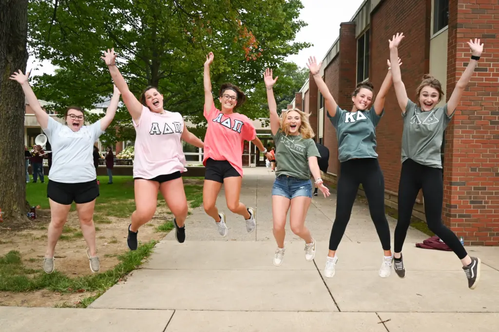 Six people are outdoors, jumping with joy in front of a brick building. Clad in casual clothing like shorts and t-shirts adorned with Greek letters, they embody the spirit of Greek Life. Greenery and a winding path add to the vibrant scene.