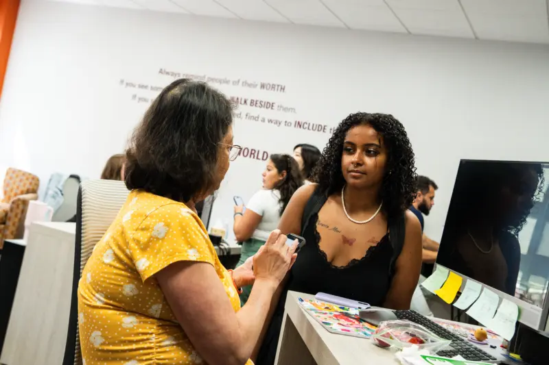In the vibrant Burke Hawk Hub, a group engages in lively conversation. A woman in a yellow shirt chats with another woman with curly hair leaning on a desk. Inspirational quotes adorn the wall behind them, while sticky notes and a computer clutter the desk's surface.