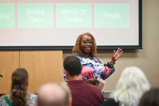 A person stands in front of an audience, speaking and gesturing with one hand. They are smiling and holding a paper. Behind them, a presentation slide with the words 