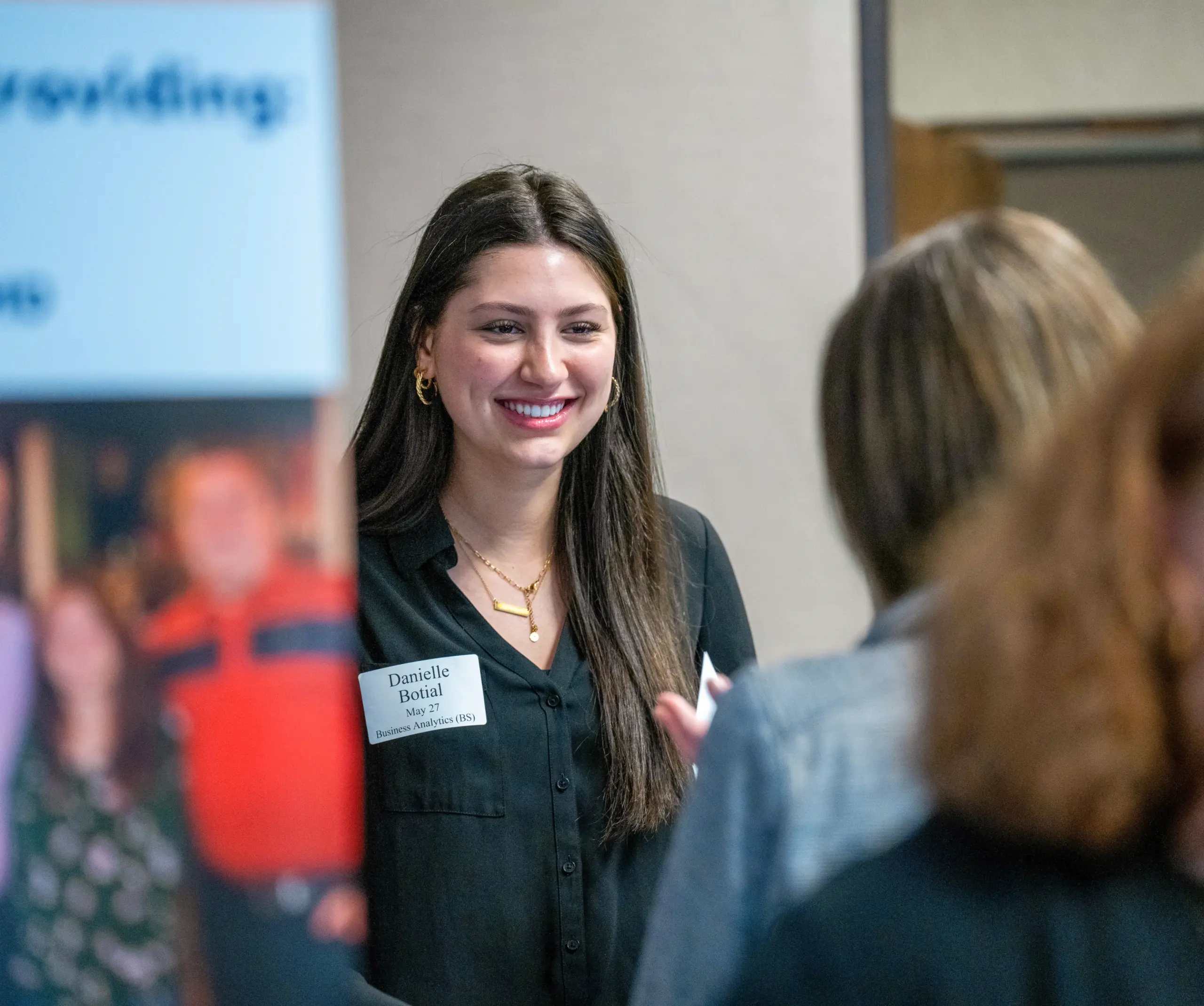 A woman with long dark hair and gold hoop earrings smiles in a professional setting, wearing a black top and name tag. She is engaged in conversation with others, standing near a blurred banner.