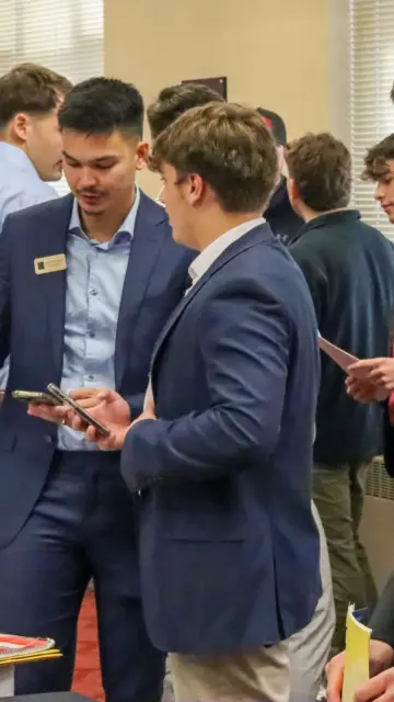 A group of people in business attire engage in conversation at a professional networking event. Some are holding papers and phones. A woman is gesturing while talking to a man. The setting is an indoor conference or meeting room.