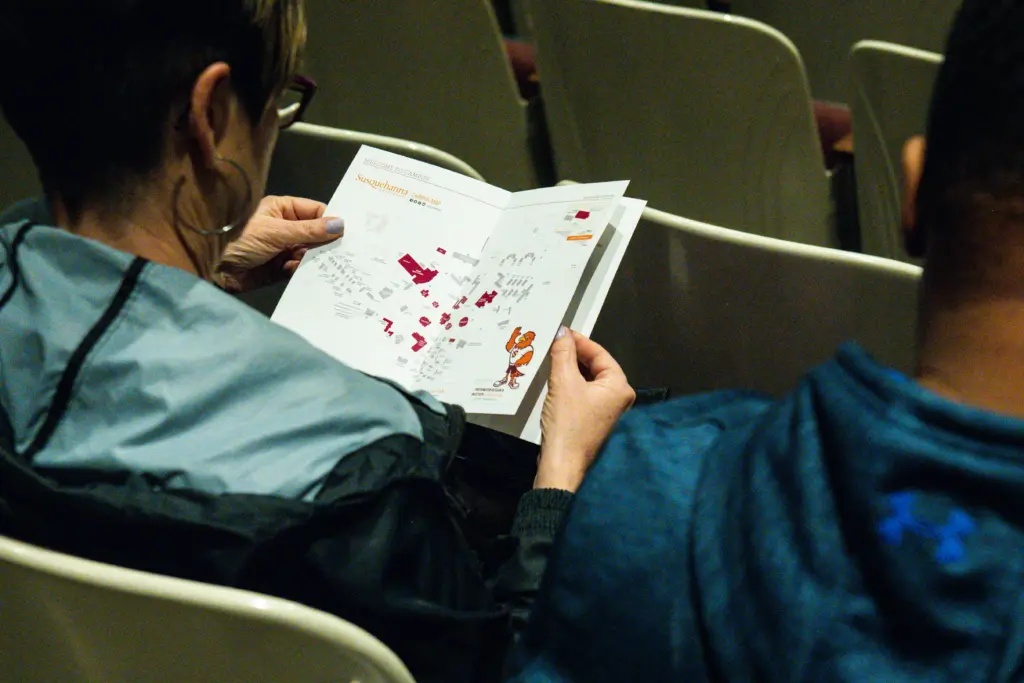 Two people sit in auditorium seats. The person on the left holds an open book with colorful maps and illustrations. The person on the right, clad in a blue jacket, glances over with interest. The setting feels relaxed and educational, inviting exploration and discovery of new directions.
