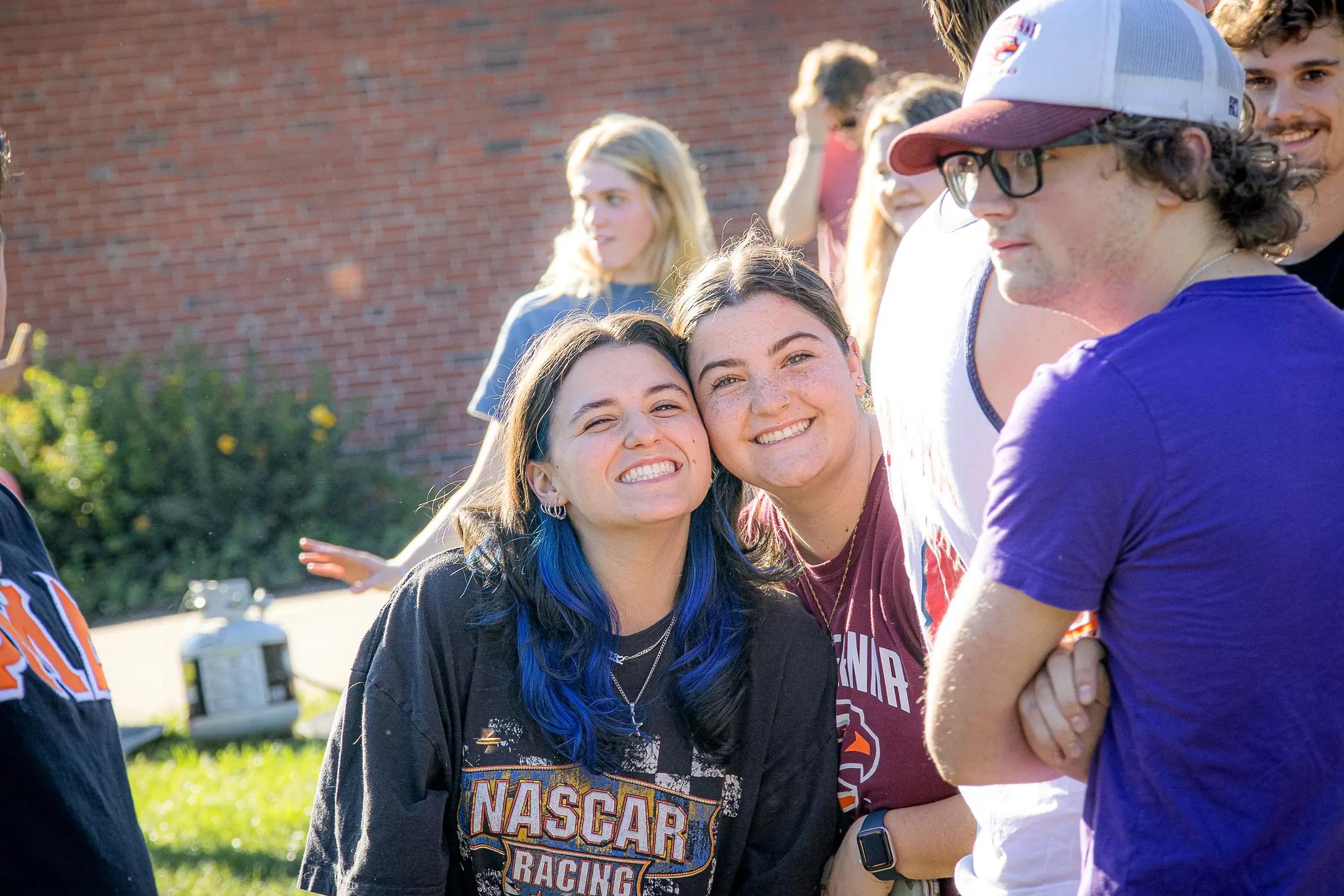 A group of young adults, embodying the spirit of Greek Life, smiles outdoors. Two in the front pose with cheerful expressions as the sun shines on a backdrop of a brick wall and grass. Casually dressed, they seem to be thoroughly enjoying the event's vibrant atmosphere.