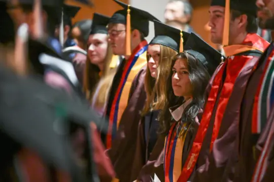 A group of university graduates in caps and gowns stand in a row during a commencement ceremony. The focus is on a female graduate in the middle, with others slightly blurred. They wear stoles and sashes, looking attentive.