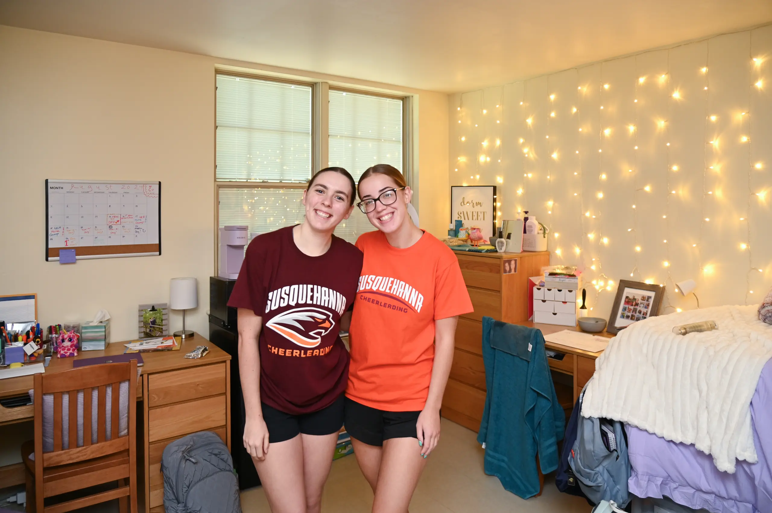 Two people stand smiling in a cozy first-year housing dorm room. Both wear 体育买球 University T-shirts, one in maroon and the other in orange. The room features a neatly organized desk, a bed, twinkling string lights, and various decorations.