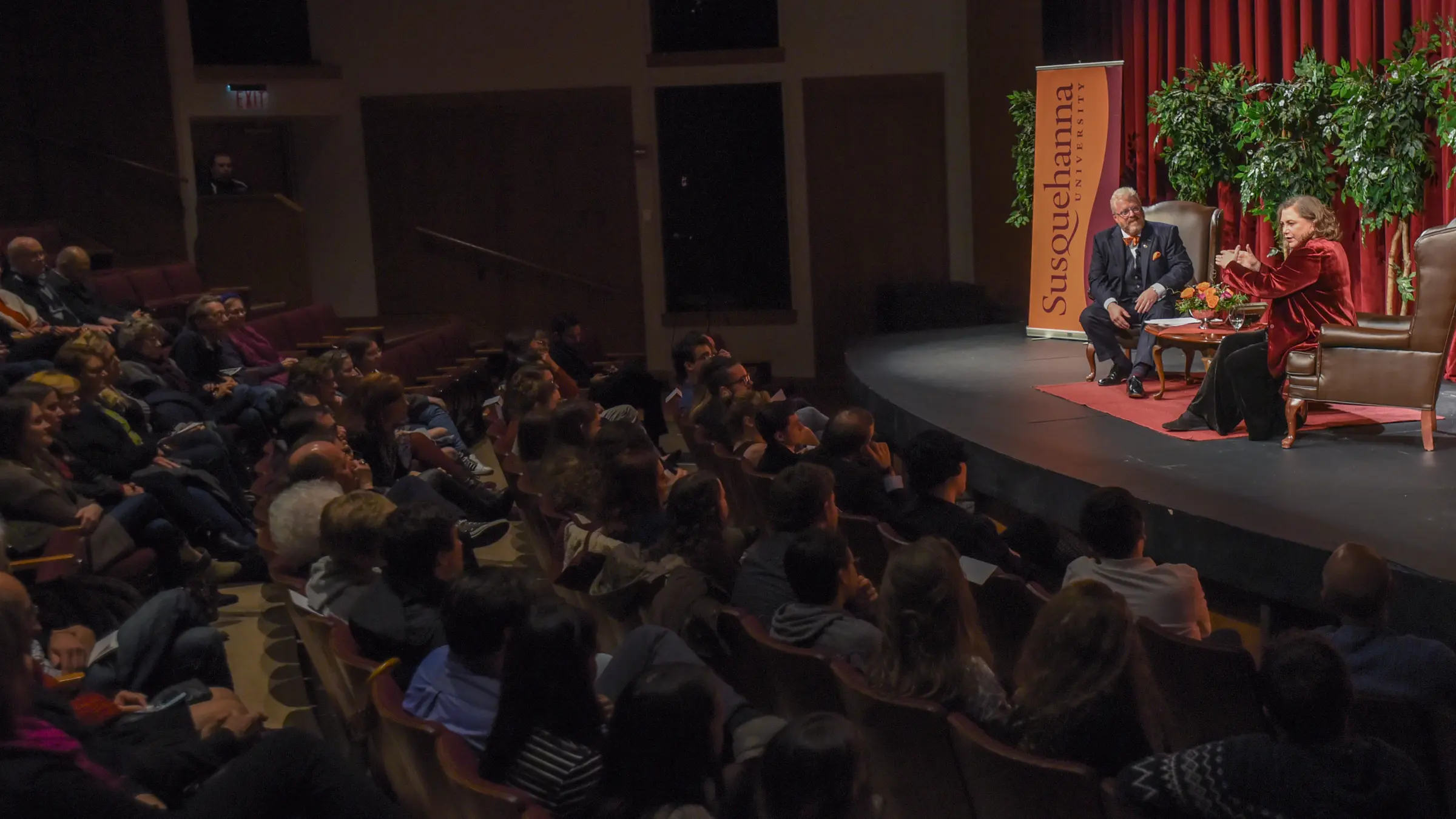 Two people are seated on stage in a dimly lit auditorium for the Dr. Bruce L. Nary Theatre Guest Artist Lecture. A red curtain and a 