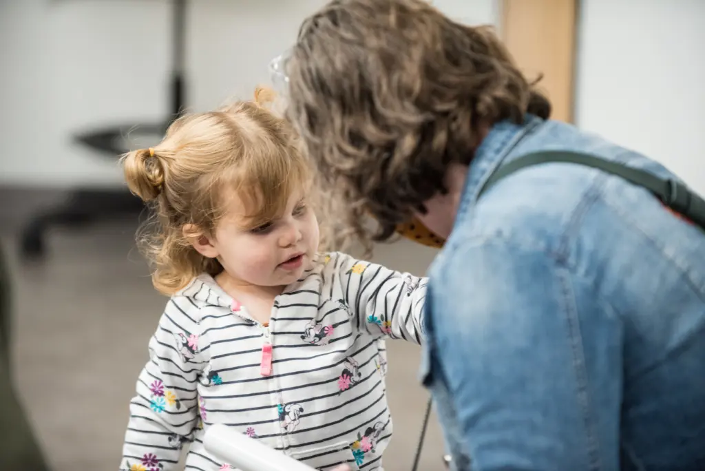 A toddler with two ponytails, wearing a striped floral shirt, touches the shoulder of an adult in a denim jacket who is crouched down to her level. They are indoors, in a softly lit room.