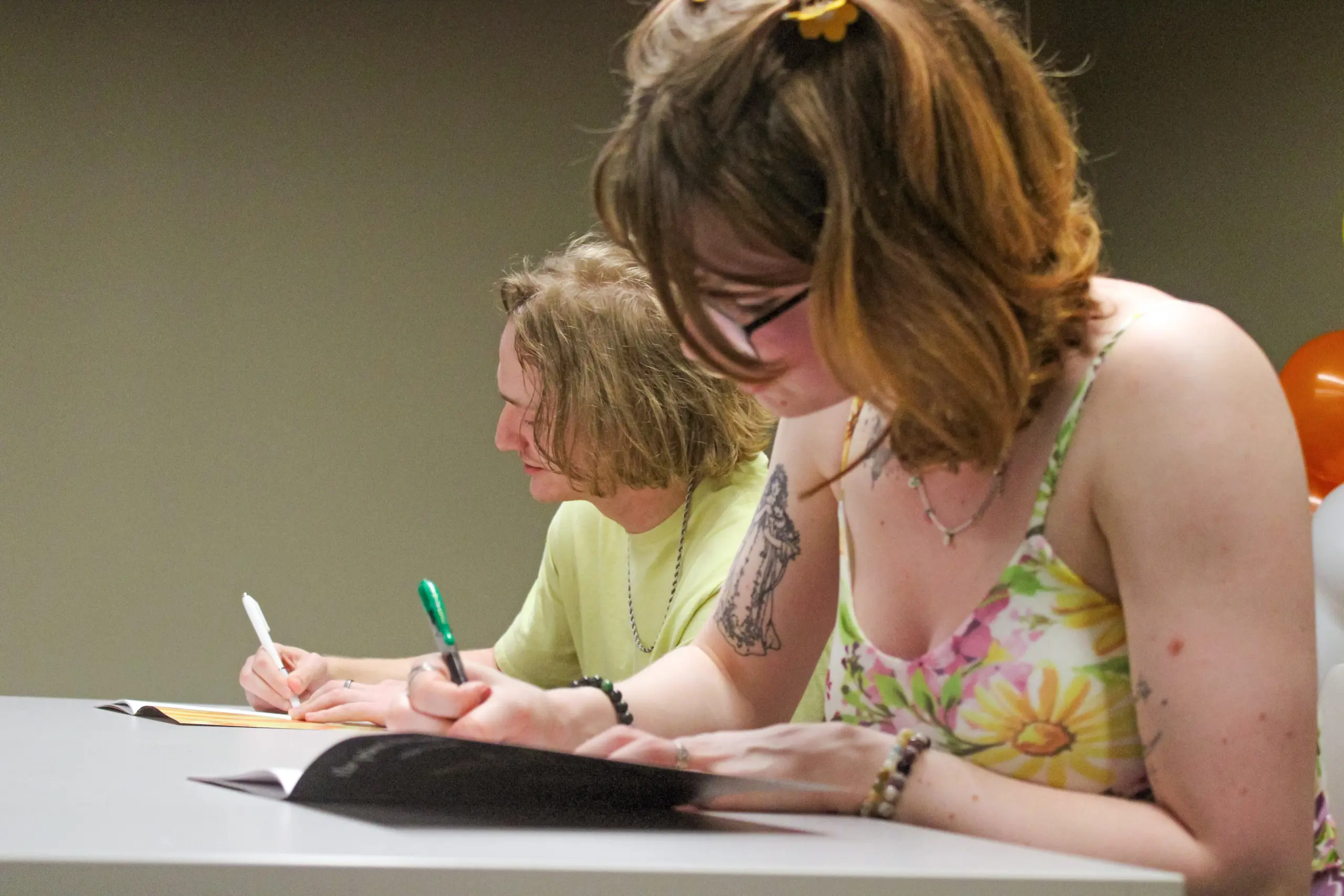 Two people are seated at a table, focused on writing in notebooks. The person in front wears glasses and a floral dress, while the person in the back wears a yellow shirt. Both have pens and there are balloons in the background.