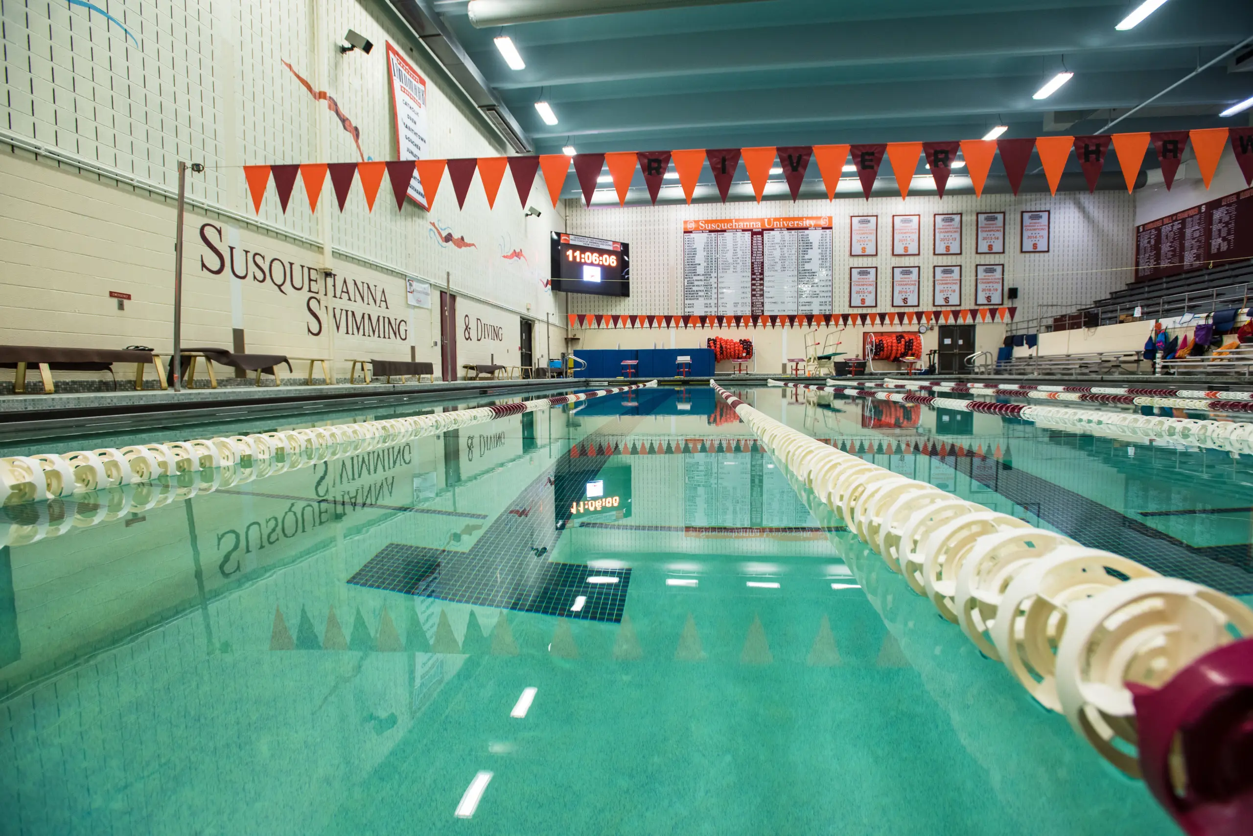 Indoor swimming pool with lane dividers, orange and black pennant banners, and a digital scoreboard on the wall. The words 