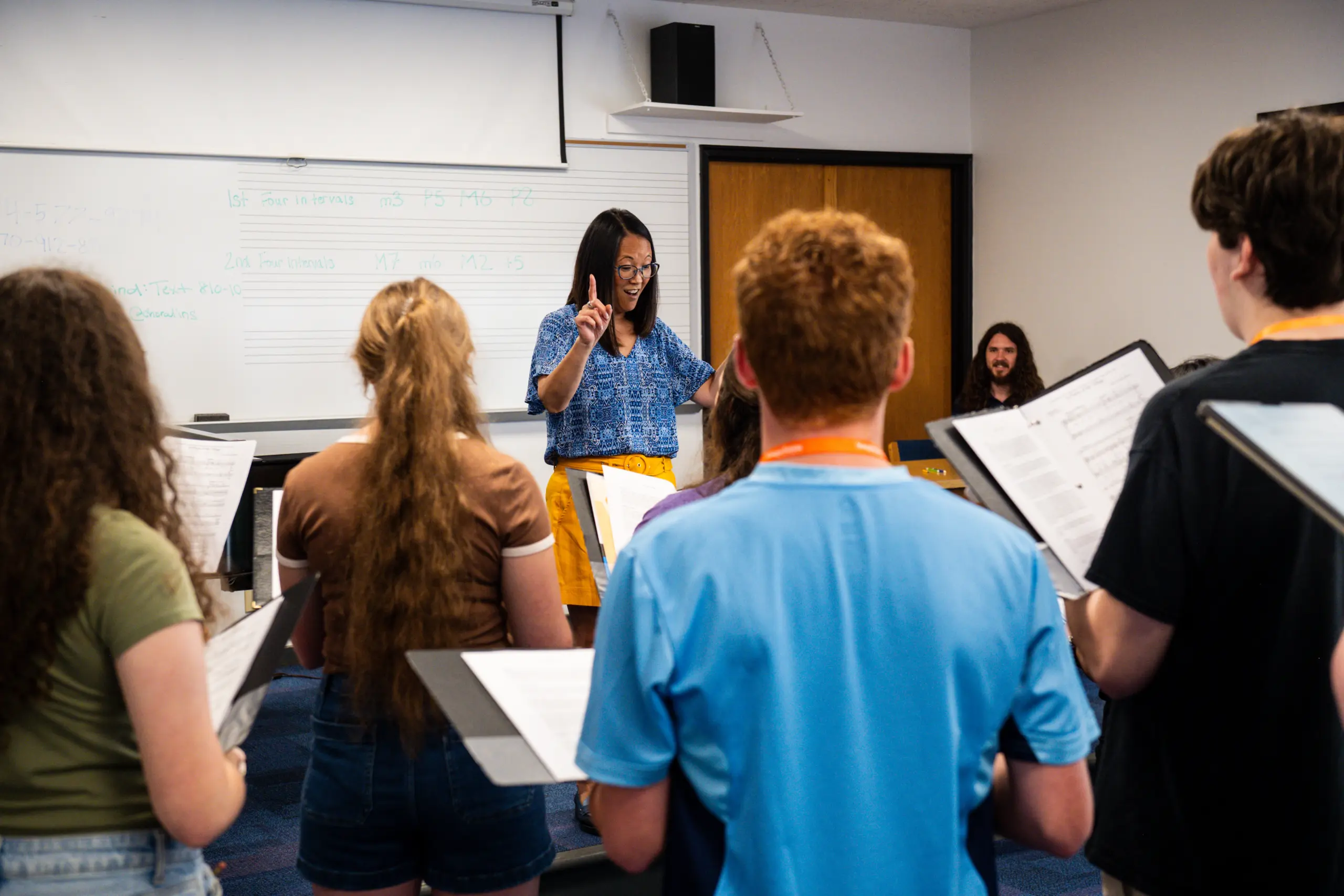 A woman conducts a group of people holding sheet music in a classroom. She stands in front of a whiteboard with musical notes. The participants face the conductor, and some wear brightly colored shirts.