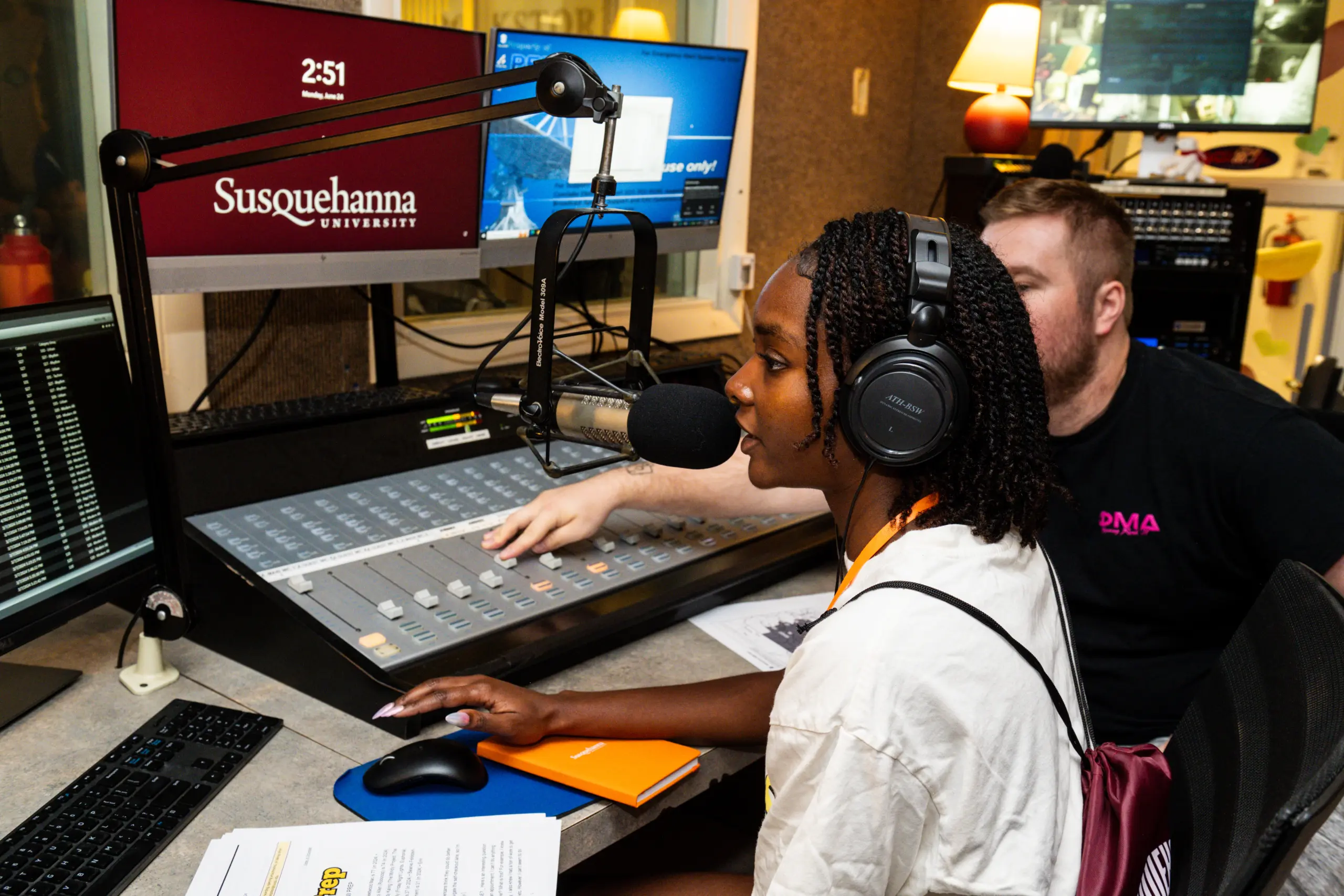 A woman speaks into a microphone in a radio studio, wearing headphones. A man sits beside her, adjusting controls on the soundboard. A computer screen displays 