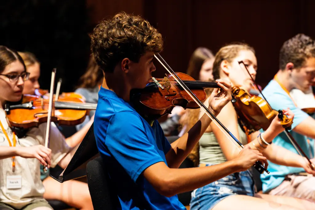Teenagers playing violins in an orchestra during a rehearsal or performance. The focus is on a young man in a blue shirt, with other musicians in the background. The setting is dimly lit, emphasizing the musicians' concentration.