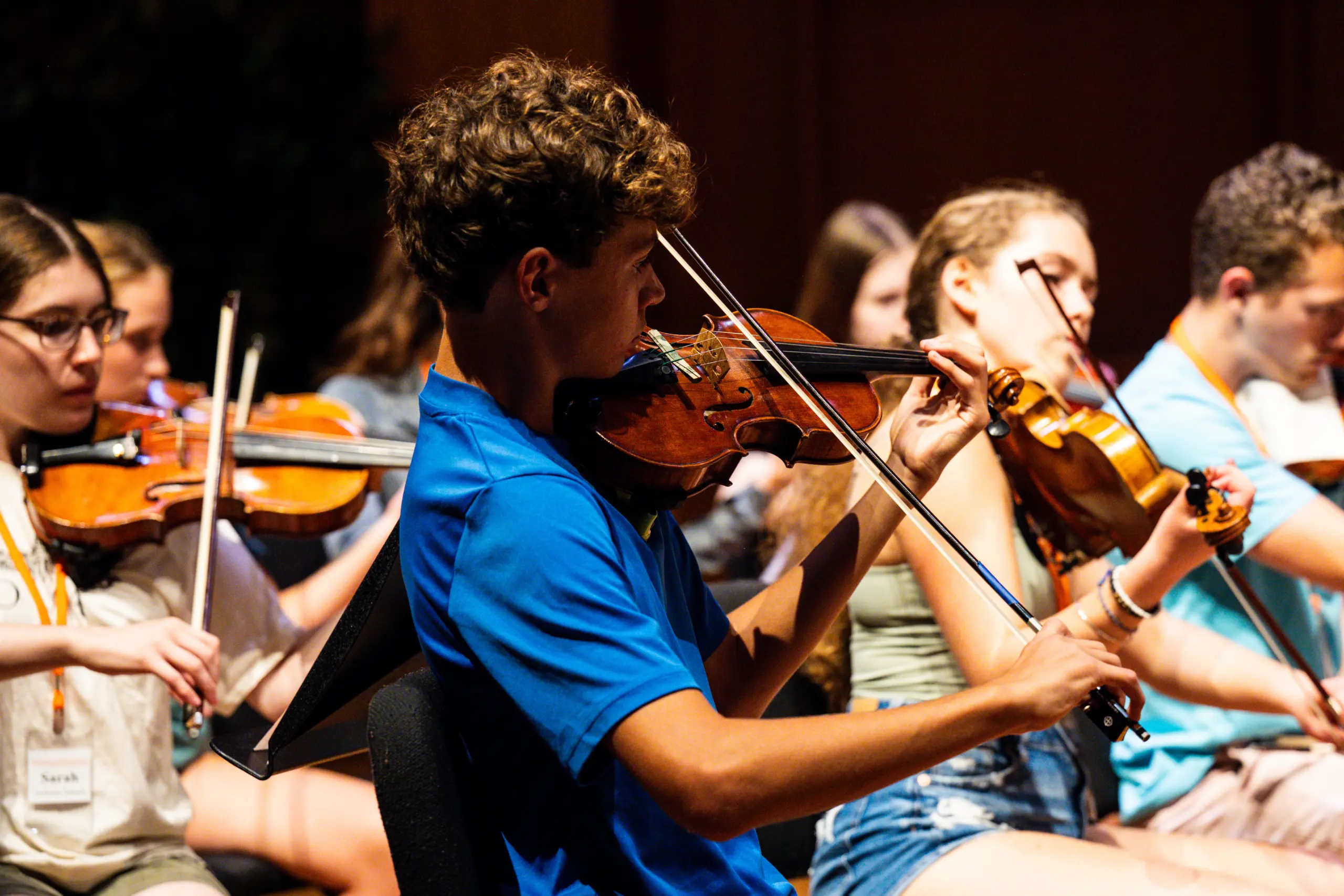 Teenagers playing violins in an orchestra during a rehearsal or performance. The focus is on a young man in a blue shirt, with other musicians in the background. The setting is dimly lit, emphasizing the musicians' concentration.