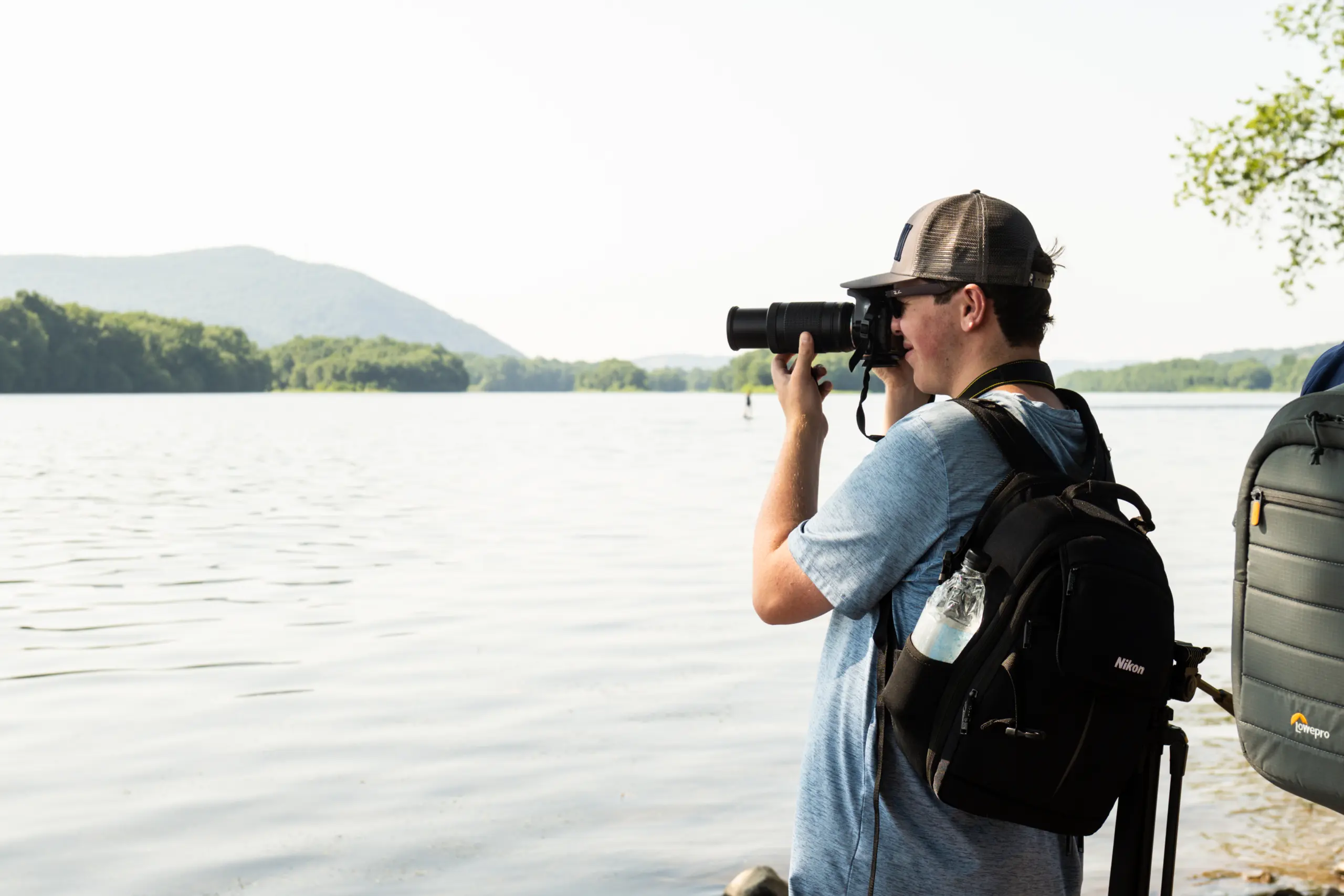 A person in a blue shirt and cap is taking a photograph with a camera near a calm, expansive body of water. They have a black backpack and a water bottle attached, with a distant view of mountains and trees under a clear sky.