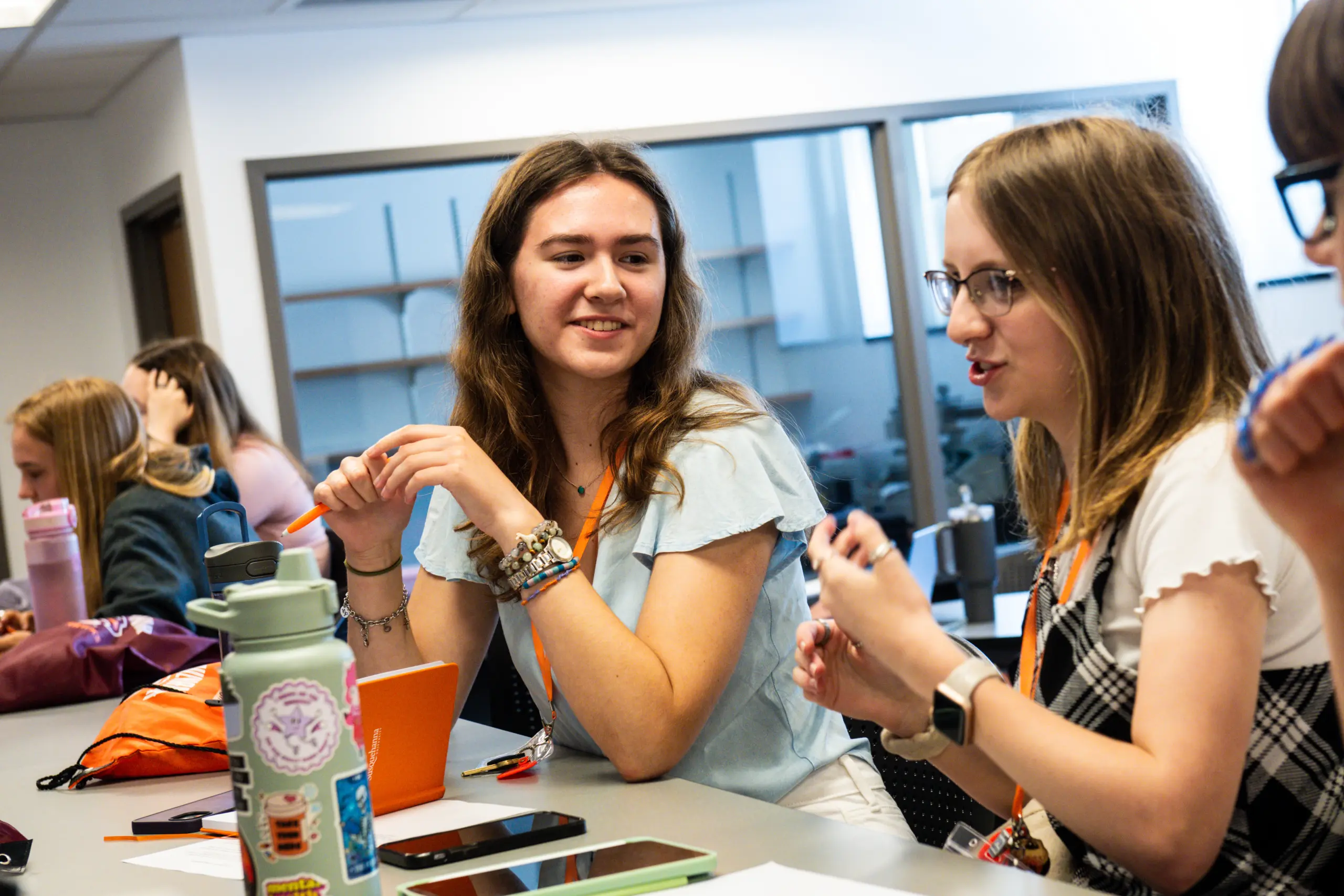 A group of young adults sitting around a table in a classroom, engaged in discussion. The focus is on two women in conversation; one with a light blue shirt, and the other with a white top and checkered dress. Tablets and water bottles are on the table.