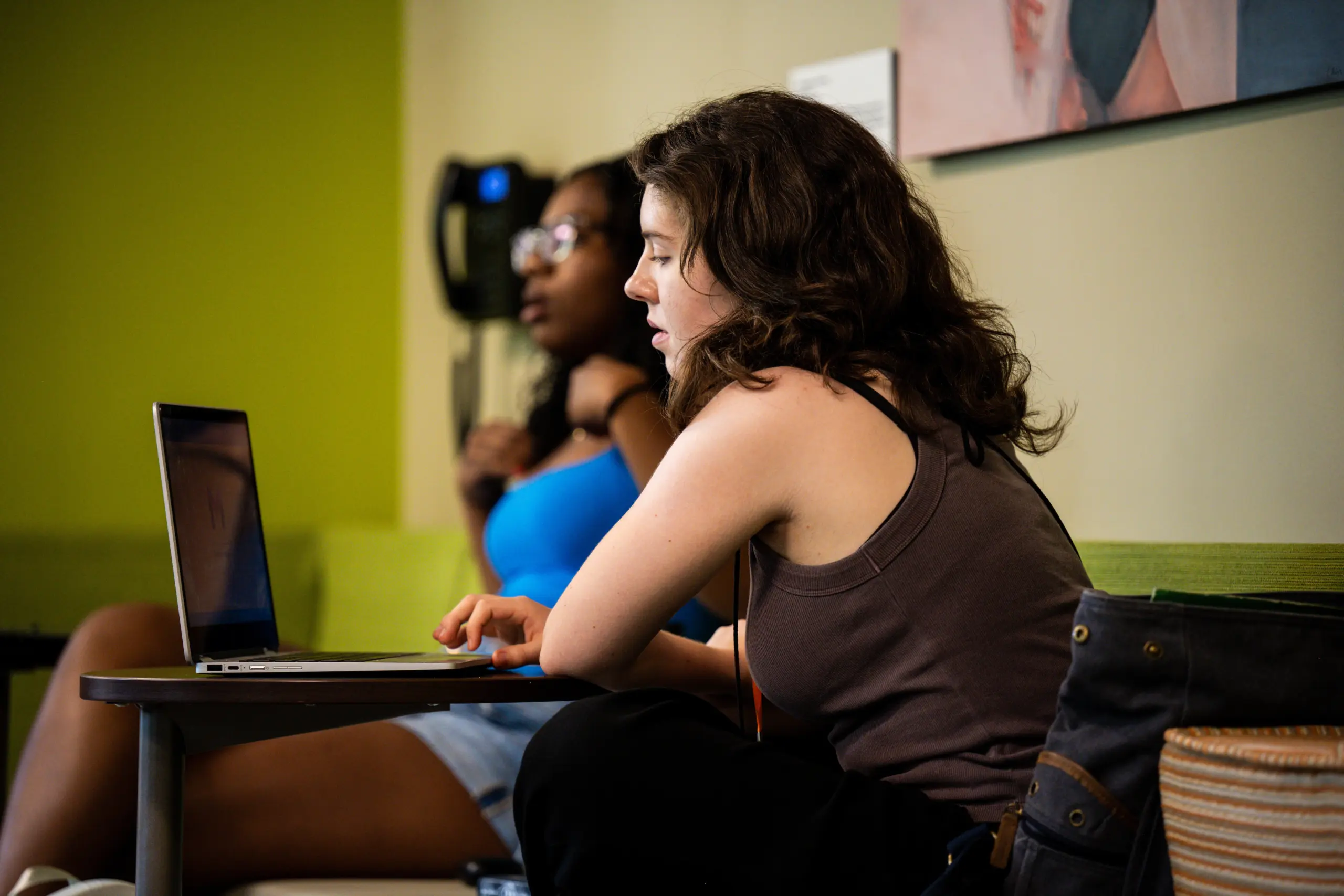 Two women sitting in a room; one in a brown tank top using a laptop on a small table, appearing focused. The other in a blue top, sitting in the background with a relaxed posture. The room has green walls and a striped pillow on a seat.