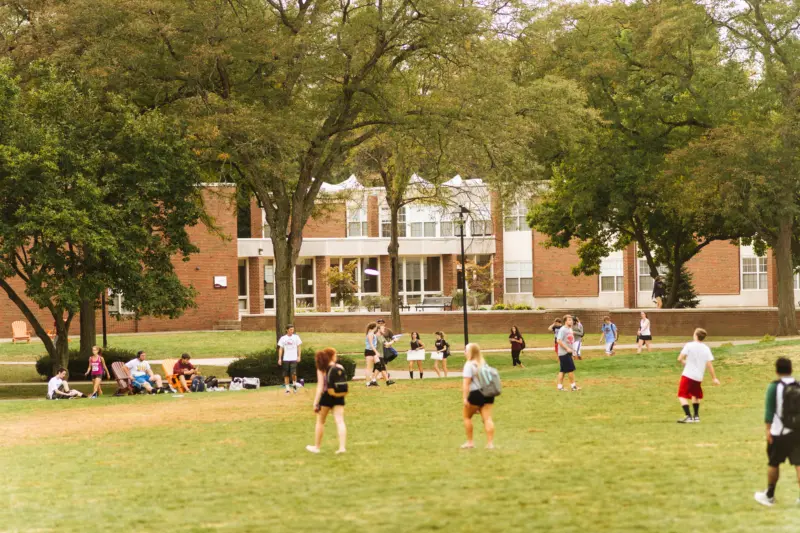 People playing soccer on a grassy field in front of a brick building with large windows. Trees surround the area, and some individuals sit on benches watching the game. It's a bright, sunny day.