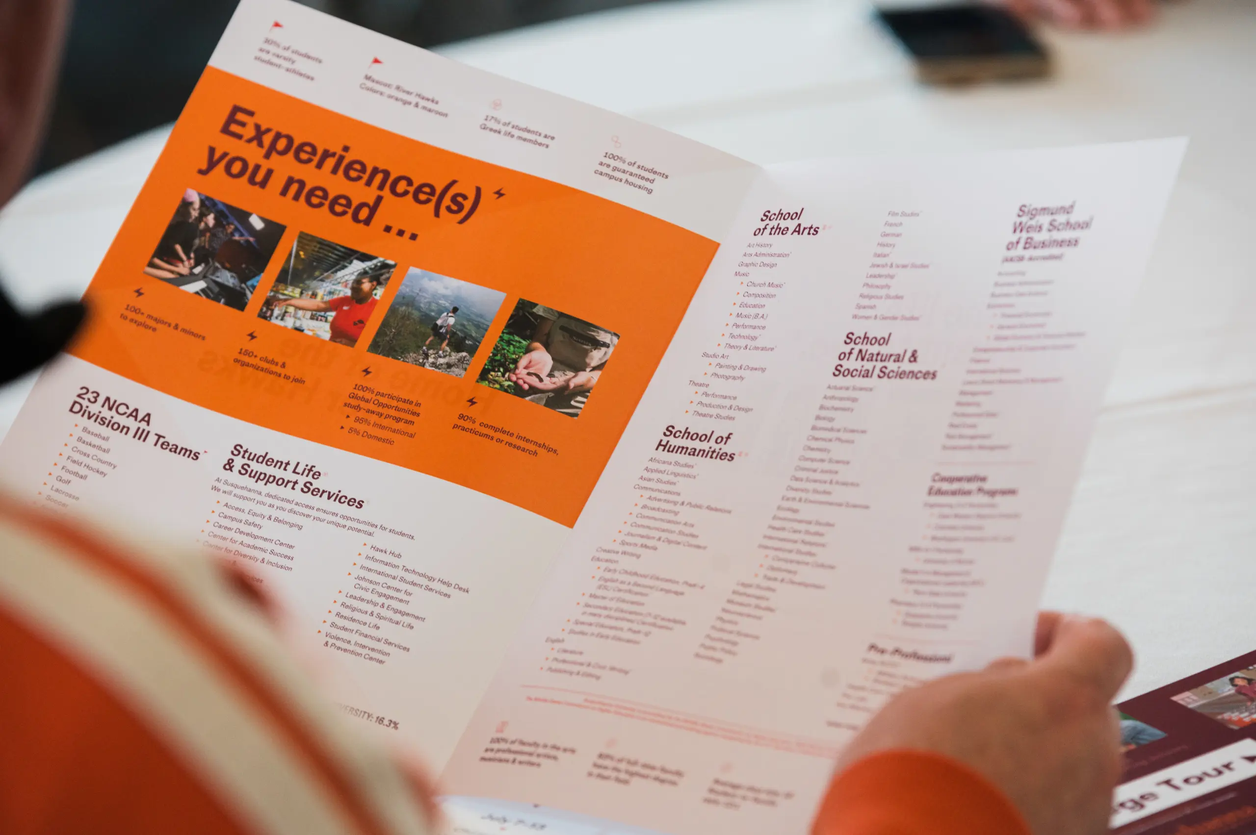 A person holds an open brochure with orange and white sections. The orange section highlights experiences, while the white section lists various schools and academic programs. The table beneath is partially visible.