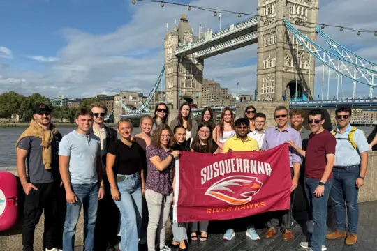 A group of people stands in front of Tower Bridge, holding a red sign with 