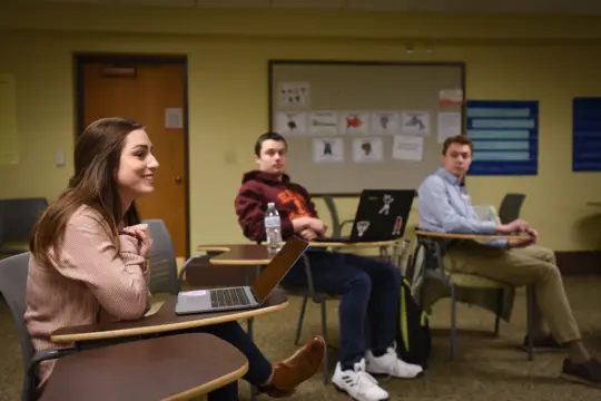 Three students sit in a classroom with desks, laptops, and a bulletin board. A woman on the left smiles while speaking. Two men, one in a maroon hoodie and the other in a blue shirt, listen attentively.