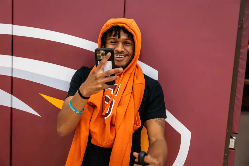 A person wearing an orange hoodie smiles while holding a smartphone and taking a selfie. They are standing against a maroon background with a white and yellow design.
