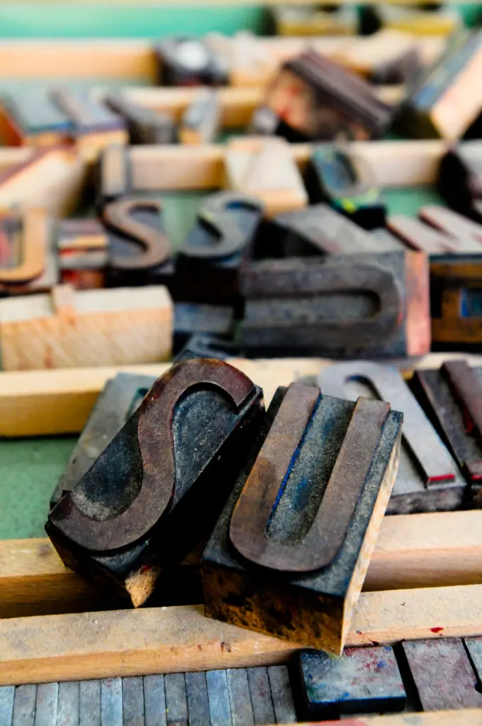 Wooden letterpress blocks scattered on a surface, focusing on large, dark blocks featuring the letter 