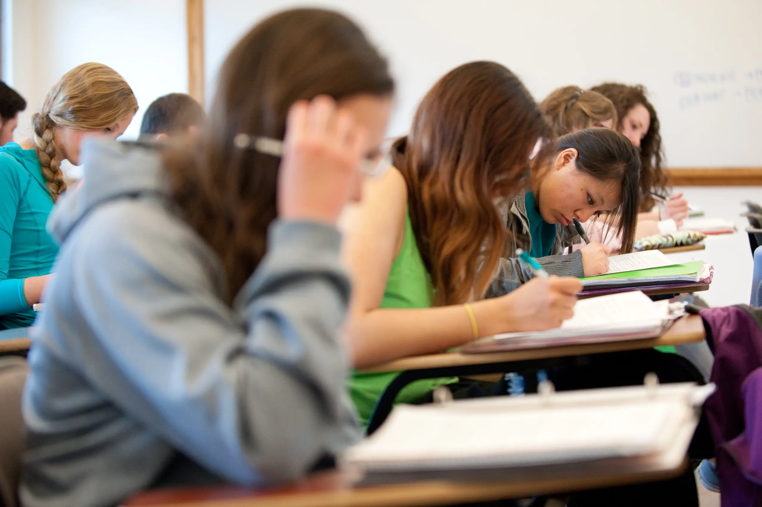 Students sitting in a classroom, focused on writing in notebooks. Some have books open, and others are wearing casual clothing such as hoodies. They are seated in rows at desks, with a whiteboard visible in the background.