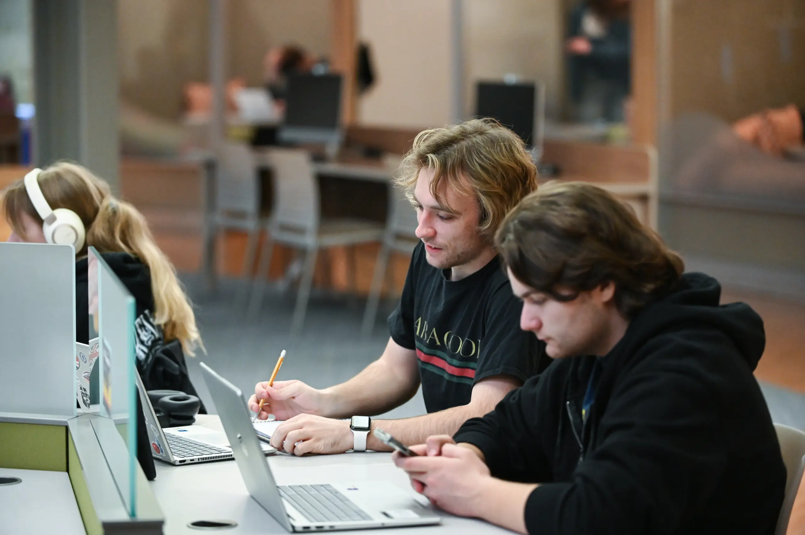 Three people sit at a table with laptops. The person in the middle has a pencil and notebook, while the other person on the right is using a smartphone. The background shows a blurred interior with chairs and tables.