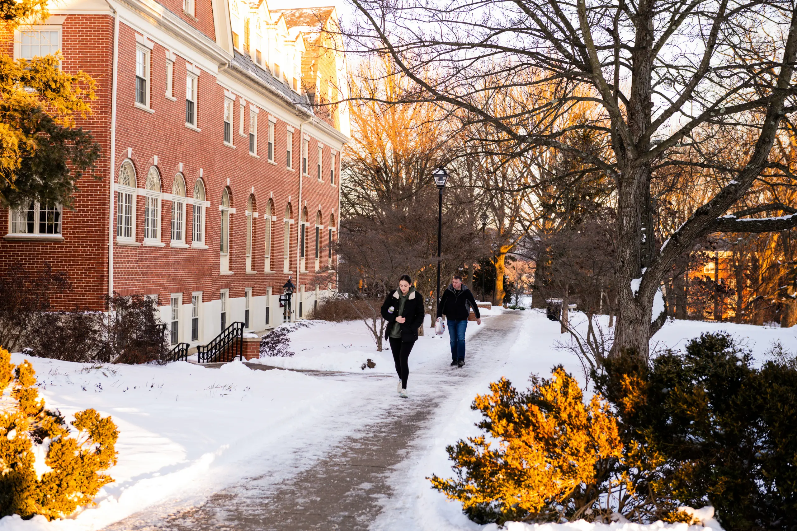 Two people walk along a snow-covered path by a brick building in winter. Bare trees and bushes line the pathway, with sunlight casting a warm glow on the scene.