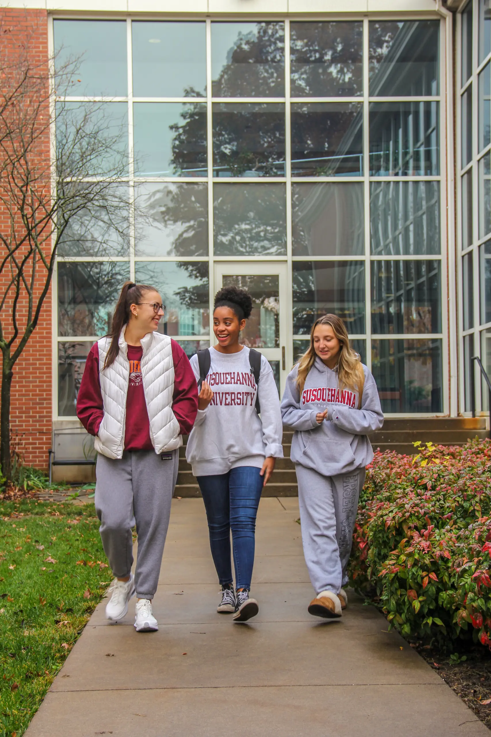 Three students walk together outside a building with large windows. They wear 体育买球官网 University apparel and carry backpacks. The path is lined with shrubs, and a tree with bare branches is visible on the grassy area.