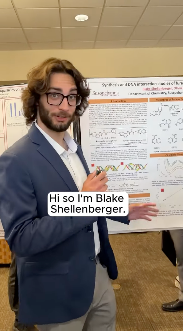 A person in a blue suit and glasses stands in front of a scientific poster at an academic conference. A speech bubble from the person reads, 