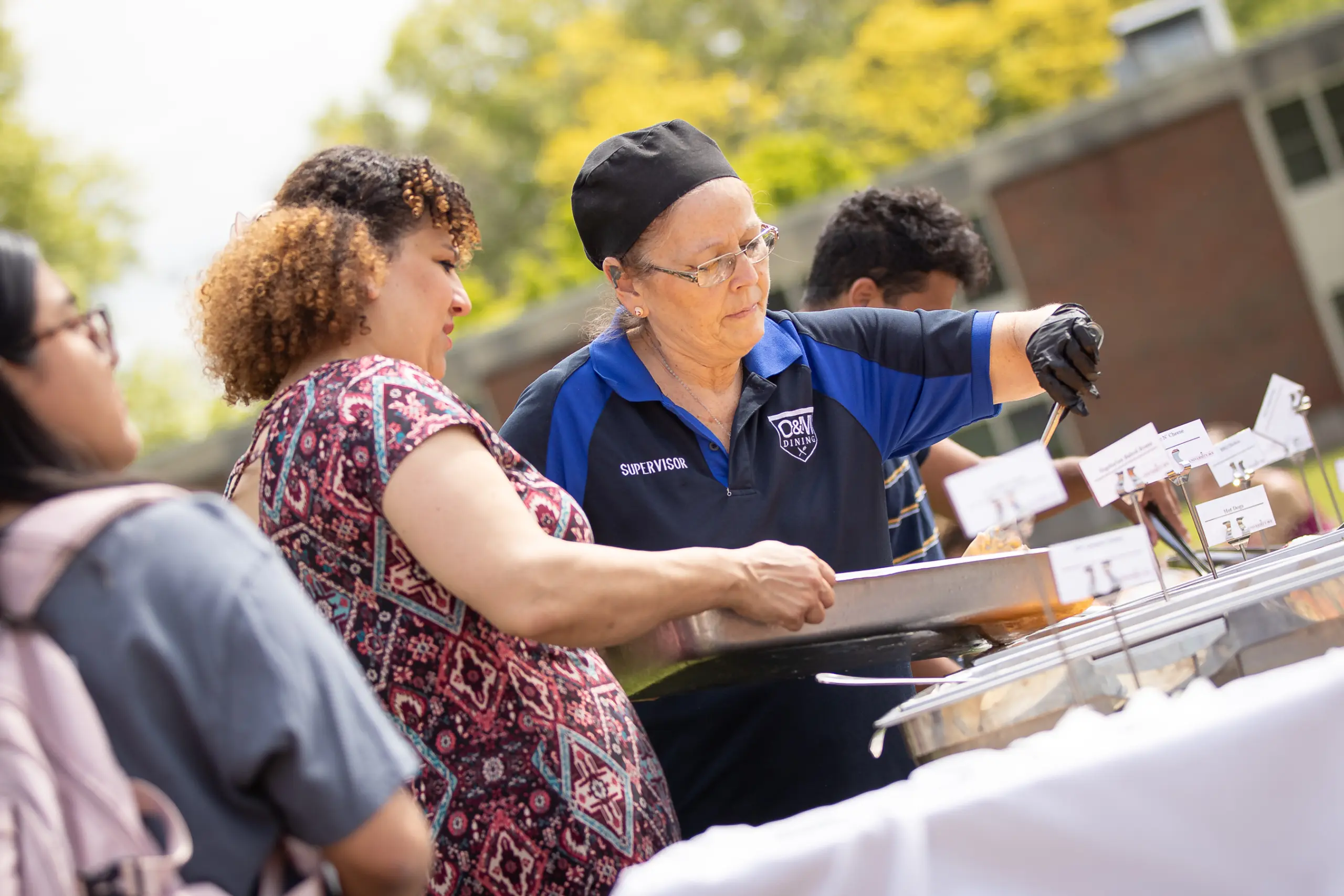 A woman in a blue polo shirt and black cap serves food from a tray at an outdoor employment event. She assists another woman holding a plate, surrounded by attendees. Tables laden with food are set up, with trees and a building providing the backdrop.