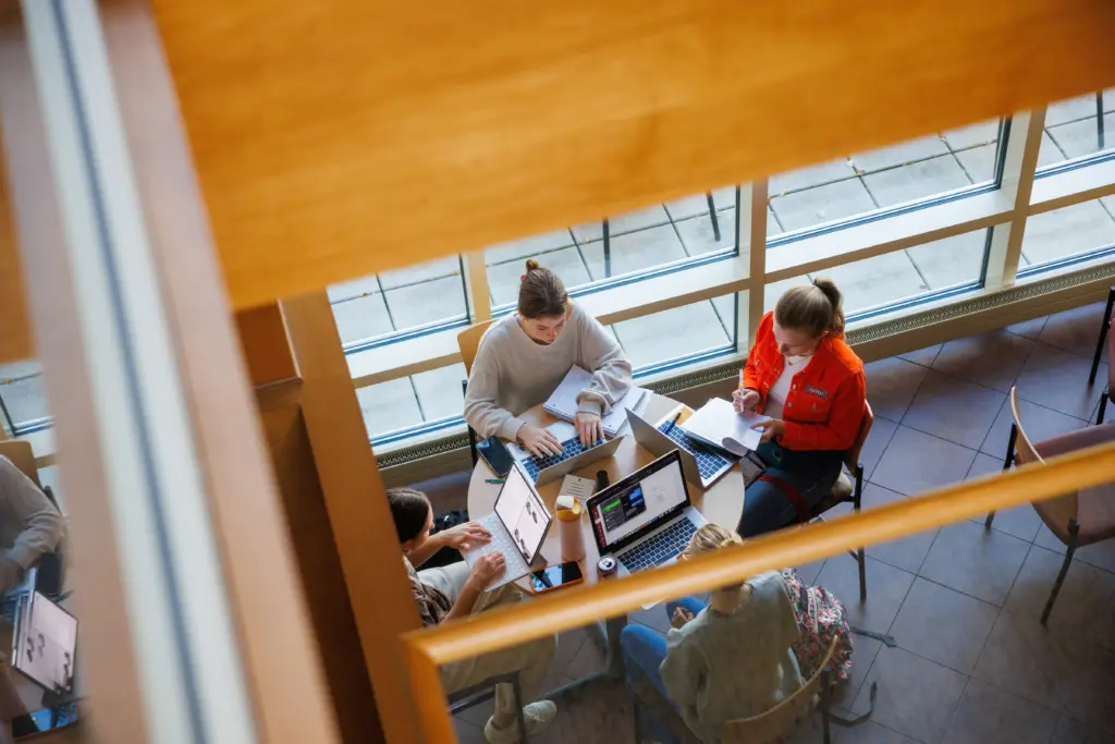 Aerial view of four people sitting around a table in a bright room with large windows. They are using laptops and notebooks, engaged in discussion. The scene suggests a collaborative study or meeting session.