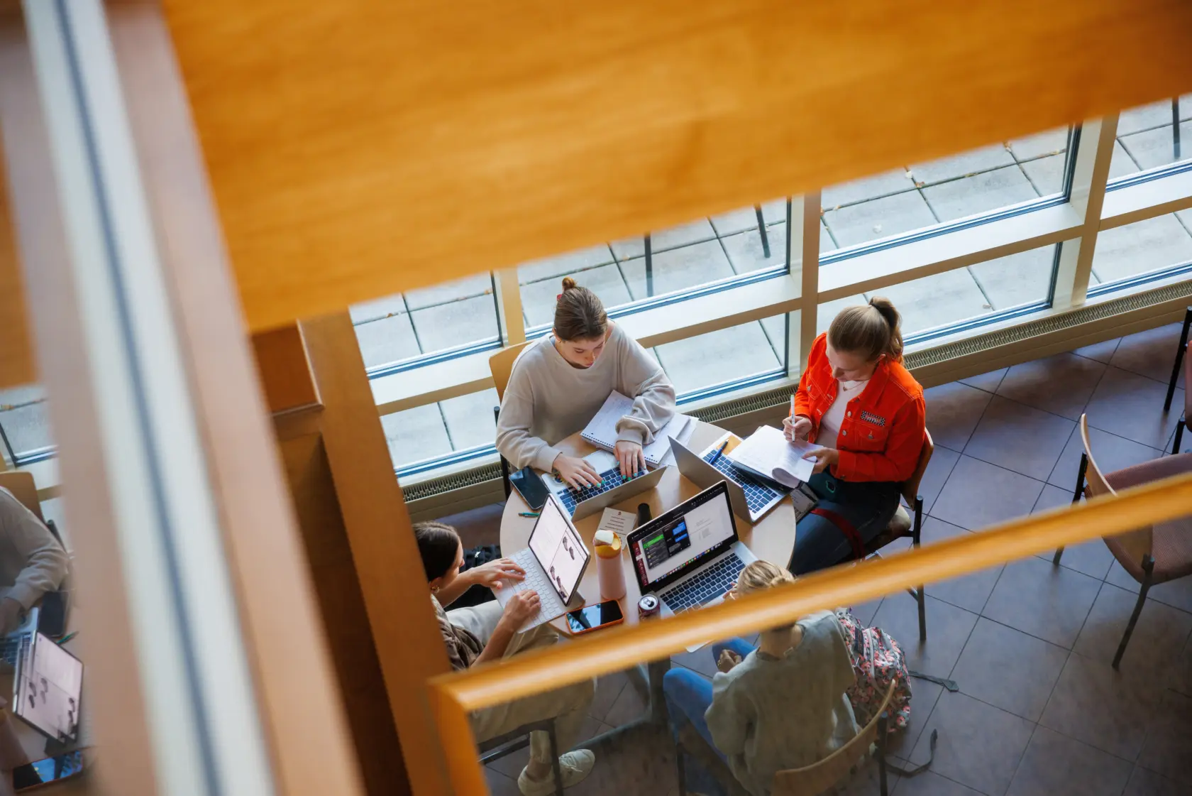 Aerial view of four people sitting around a table in a bright room with large windows. They are using laptops and notebooks, engaged in discussion. The scene suggests a collaborative study or meeting session.
