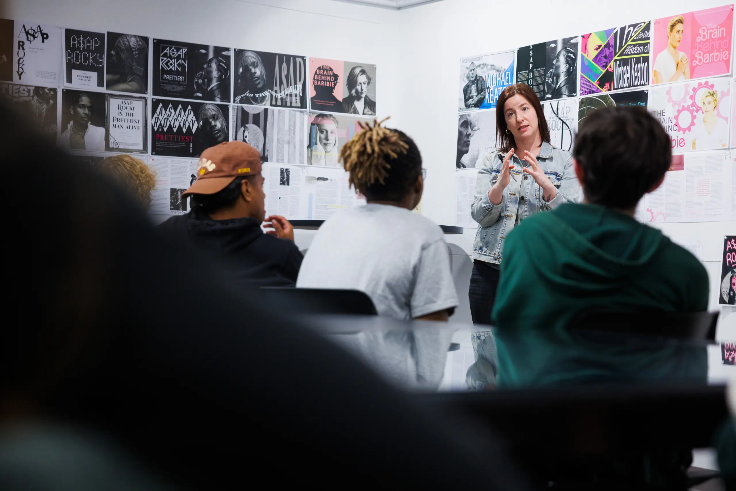 A woman stands in front of a group of seated people, gesturing as she speaks. The walls are covered with various posters and images. The focus is on her interaction with the audience in a casual, creative setting.