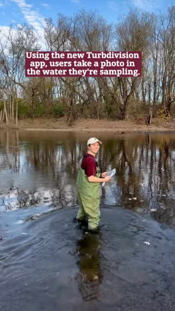A person in waders stands in a river, holding a smartphone, capturing their sampling spot. Leafless trees form the backdrop as the text highlights how the new Turbdivision app opens up exciting research opportunities by allowing users to document precisely where they work.