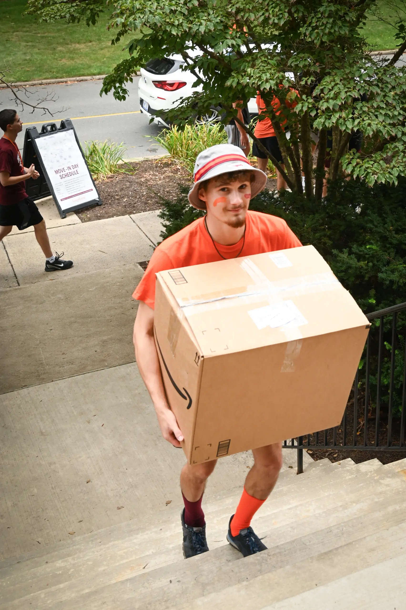 A person in an orange shirt and bucket hat is walking up outdoor steps, smiling through face paint as they carry a large cardboard box—perhaps part of their packing for college checklist. In the background, another individual is near a path surrounded by greenery.