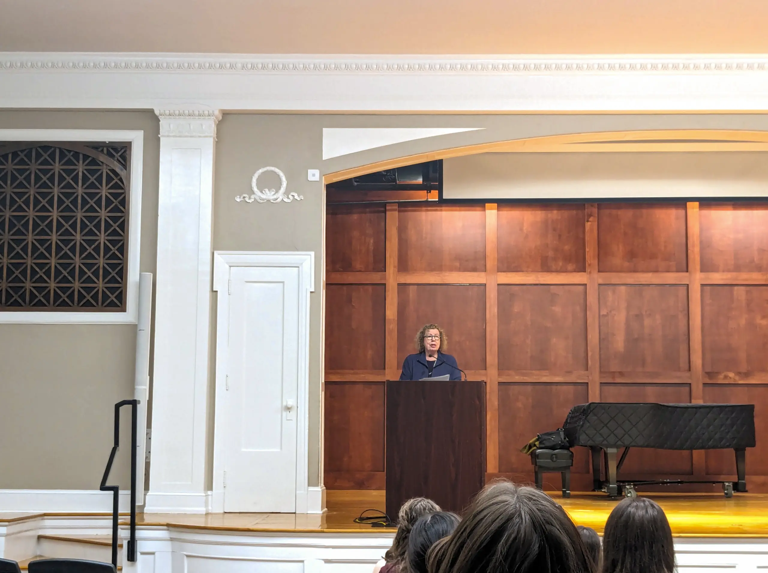 A person stands at a podium in a grand lecture hall, delivering an engaging Publishing & Editing Lecture. The room is elegantly adorned with wood paneling and white trim, while a grand piano adds sophistication. Attentive listeners fill the seats, capturing the essence of the event.
