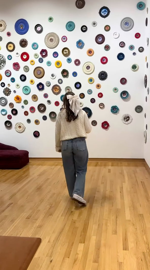 A person in a sweater and jeans walks toward a wall adorned with colorful, round crochet patterns, reminiscent of the School of the Arts' vibrant creativity. The wooden floor and a maroon sofa enhance the room's artistic ambiance.
