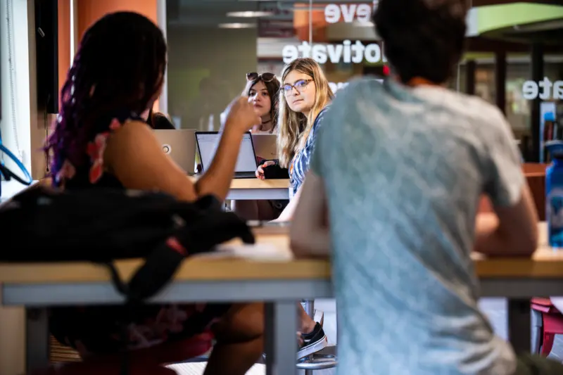 Four people, possibly international students, sit around a table in a modern room with glass walls. Two women with laptops face each other, one with glasses looking directly ahead. Two others have their backs to the camera. The room appears to be a study or meeting area.
