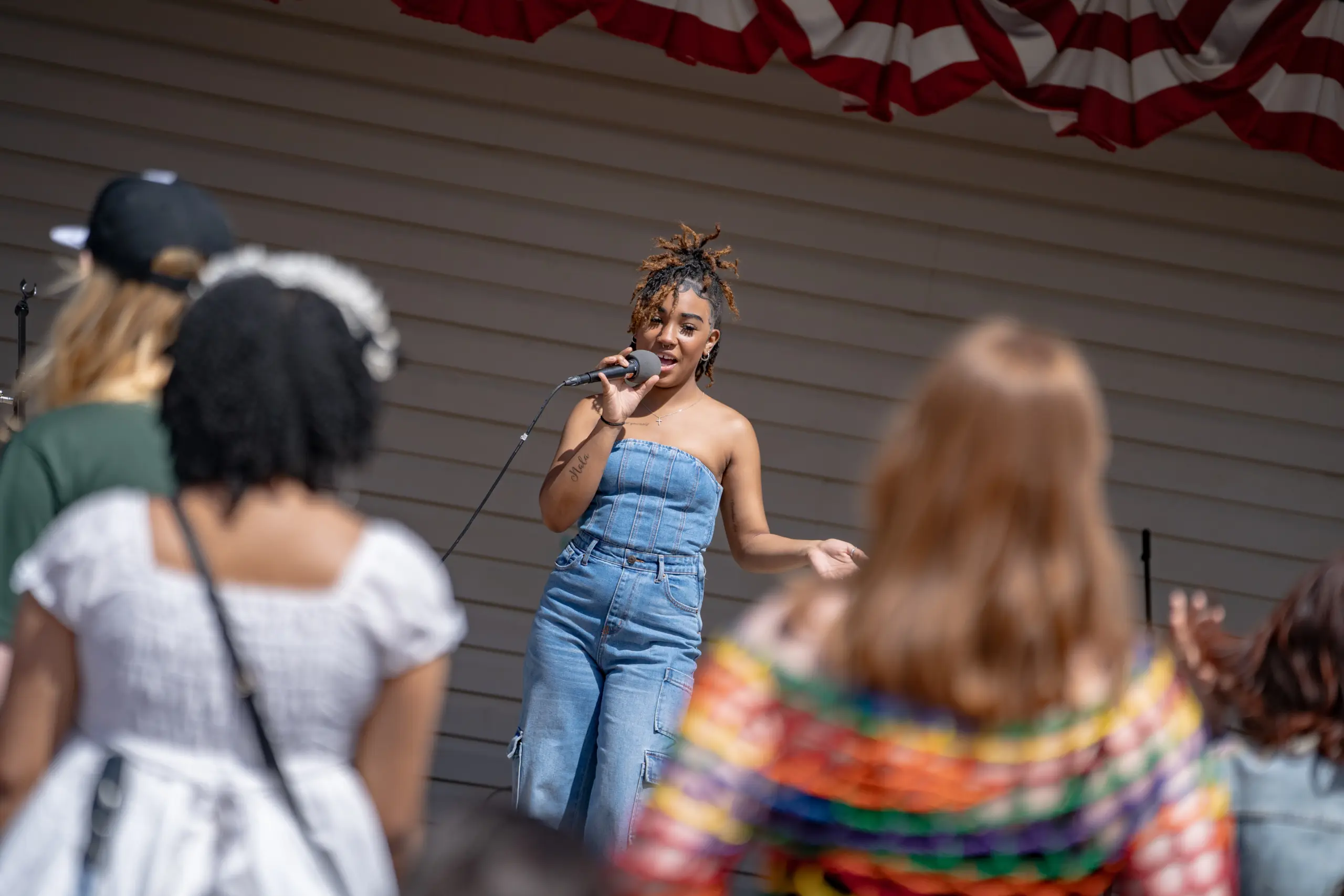 A woman in a denim jumpsuit takes the stage with a microphone, capturing the essence of campus life. The small crowd, dressed in colorful casual attire, gathers as an American flag decor adorns the scene.