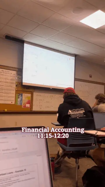 In a classroom at the Sigmund Weis School of Business, students are seated facing a projector displaying a spreadsheet. The chalkboard behind them features various formulas, and text reads 