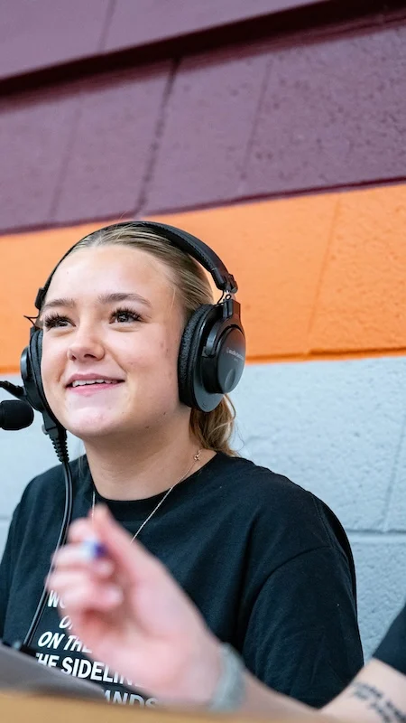 Three people are seated at a table with audio equipment. The person in the center is smiling and wearing a headset. The two others, also wearing headsets, are engaged in conversation. The background features a two-tone wall with maroon and orange stripes.