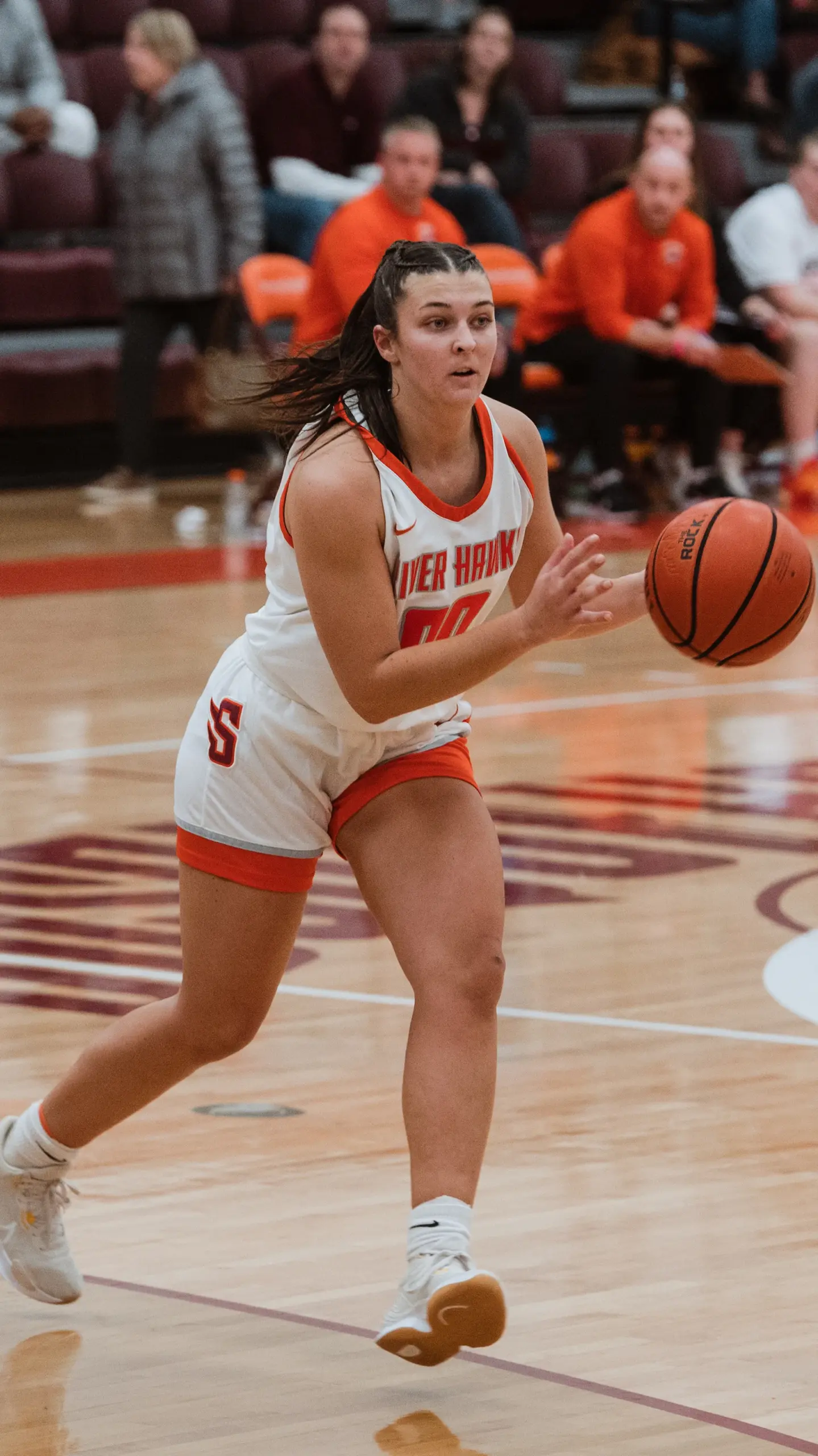 A female basketball player in a white and red uniform skillfully dribbles the ball down the court, showcasing her prowess in athletics, pursued by an opponent clad in black and blue. Teammates and spectators watch intently from the gymnasium sidelines.