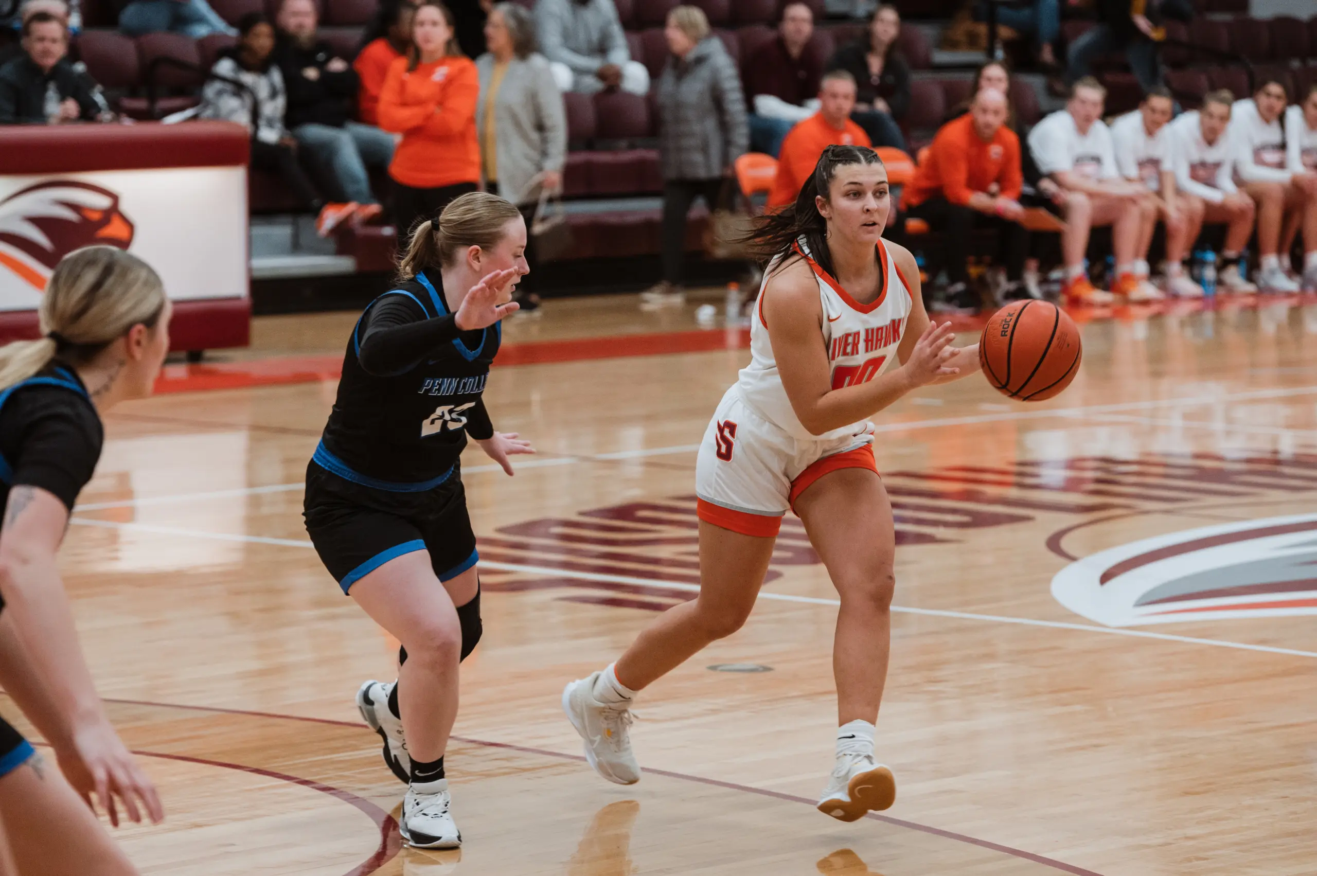 A female basketball player in a white and red uniform skillfully dribbles the ball down the court, showcasing her prowess in athletics, pursued by an opponent clad in black and blue. Teammates and spectators watch intently from the gymnasium sidelines.