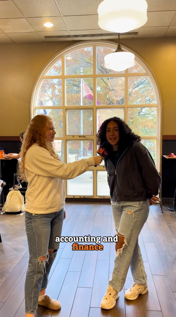 A person with curly hair is speaking into a microphone held by another person with long hair. They are indoors, standing in front of a large arched window with trees visible outside. The text reads, 