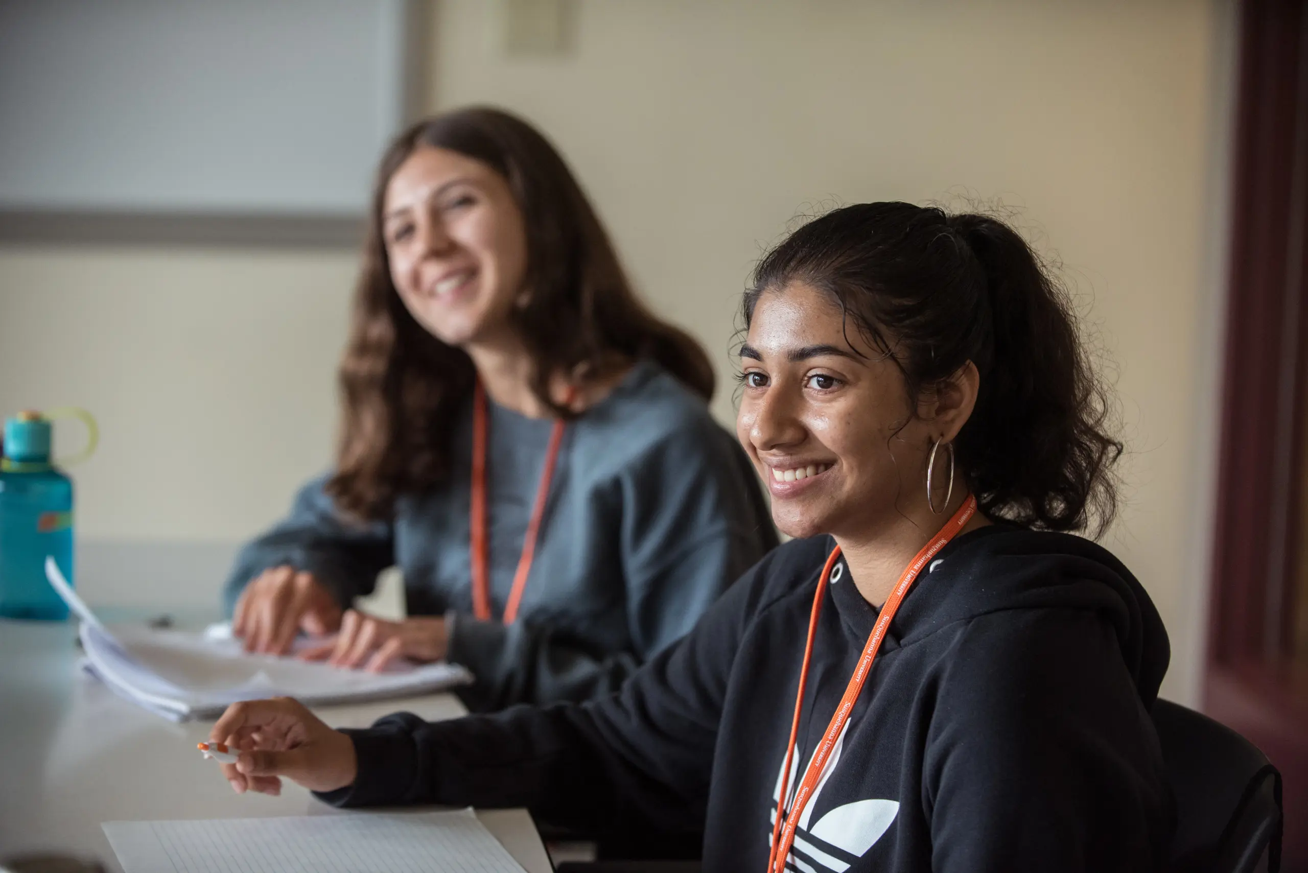Two people sit at a table in a classroom, engaged in an activity. One is writing in a notebook while smiling. Both are wearing lanyards. There is a bottle on the table, and a whiteboard in the background.