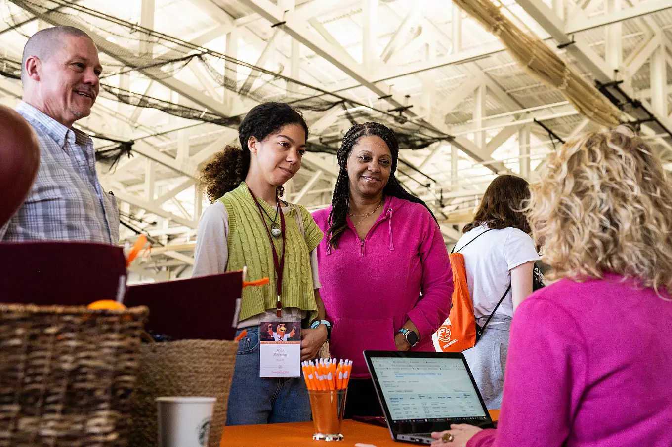 A group of three people stand at a table talking to a fourth person who is seated. The setting appears to be indoors with a high ceiling and metal beams. A laptop and basket are on the table, along with pens and an orange cloth.