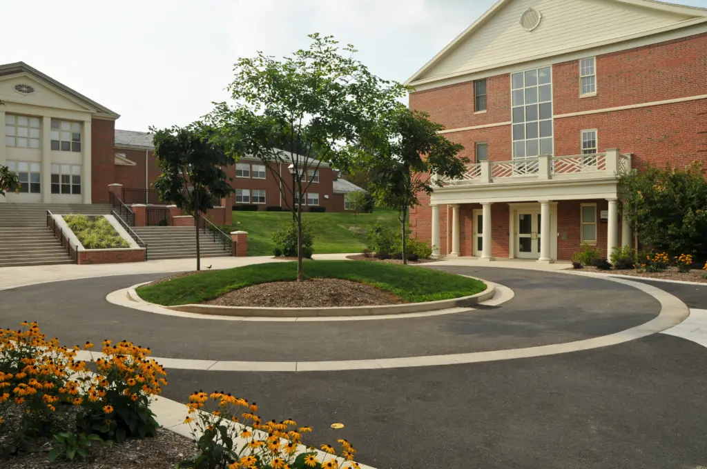 A circular driveway with a small tree at its heart is surrounded by a red brick building, stairs leading to another structure, and vibrant orange flowers. This harmonious blend of nature and architecture reflects sustainability initiatives, with a lush grassy hill in the background.
