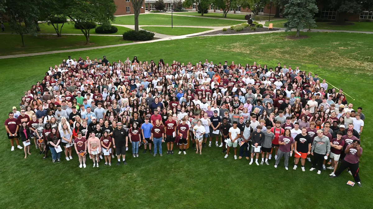 About 100 young adults stand closely together on a grassy campus area, posing for a group photo surrounded by trees and walkways.